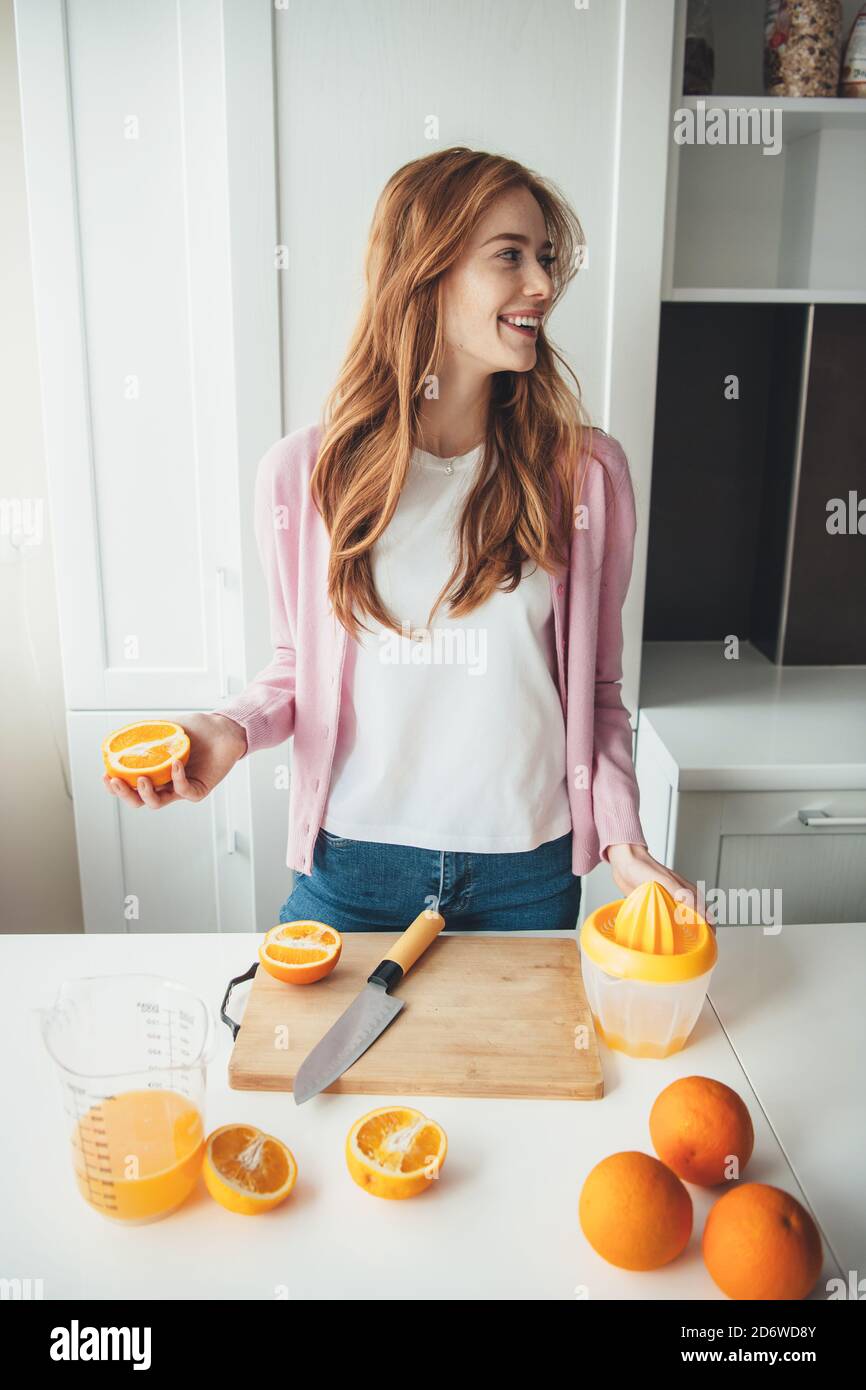 Belle femme au gingembre avec des taches de rousseur sourit tout en coupant des oranges pour préparer du jus à l'aide d'un presse-fruits manuel Banque D'Images