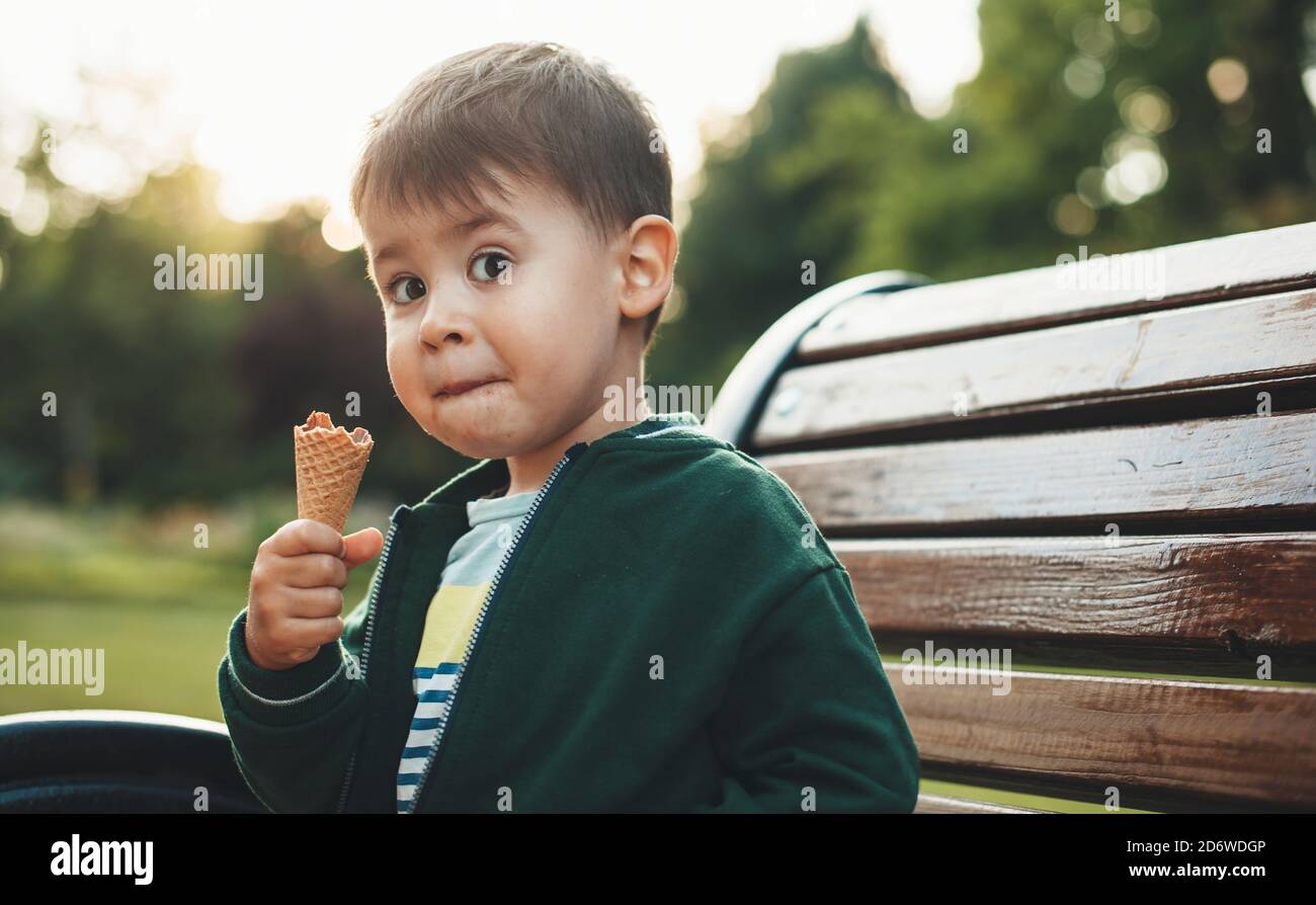 Le mignon garçon mange de la crème glacée et a l'air surpris caméra en position assise sur la banquette dans le parc Banque D'Images