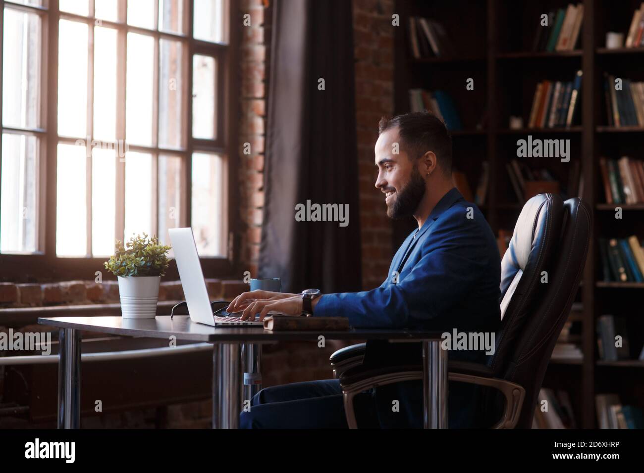 Un jeune homme d'affaires travaille sur un ordinateur portable dans un bureau luxueux de la bibliothèque. Un homme barbu, gai dans un costume bleu à un bureau est de taper sur un clavier Banque D'Images