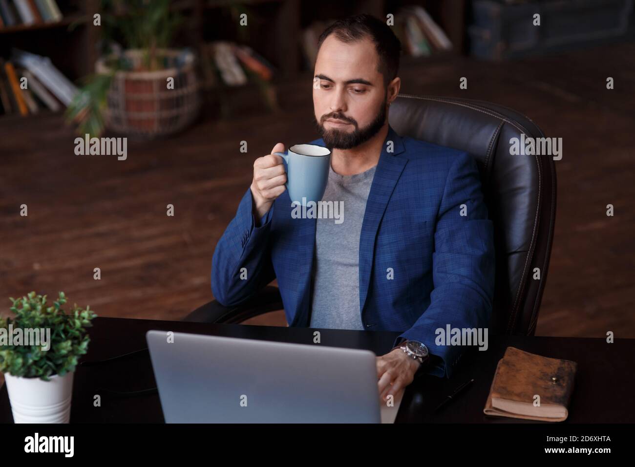 Savourez un café frais au travail. Un jeune homme plein de confiance dans un vêtement élégant et décontracté travaillant sur un ordinateur portable et buvant une boisson chaude tout en étant assis au travail Banque D'Images