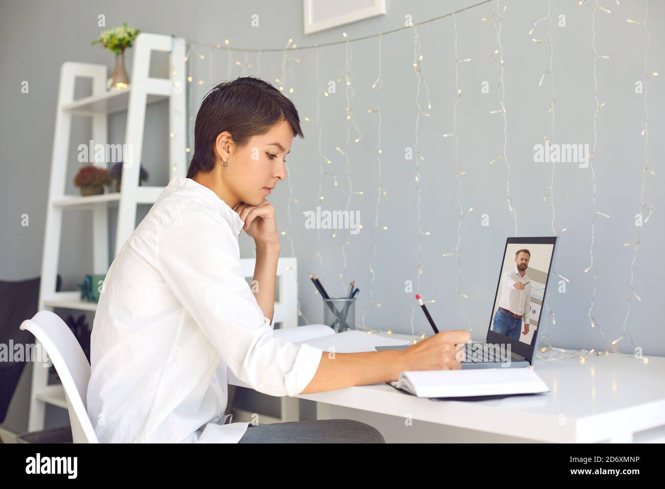 Jeune femme assise à un bureau à la maison, regardant des vidéos professionnelles éducatives sur un ordinateur portable et prenant des notes Banque D'Images