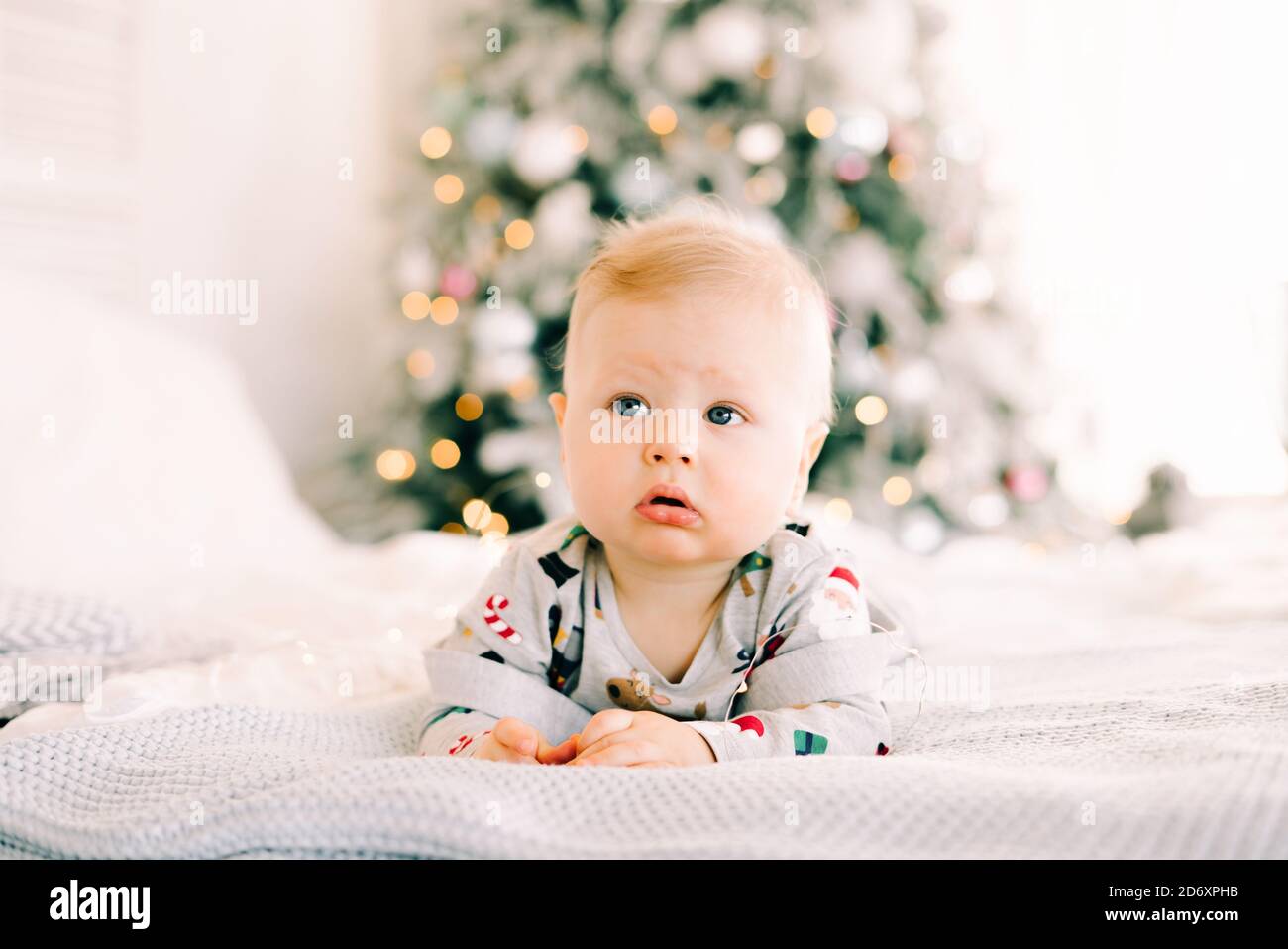 Le premier Noël de l'enfant. Un petit garçon triste et pensif est allongé dans un costume de fête sur le lit sur le fond d'un sapin aux couleurs vives Banque D'Images