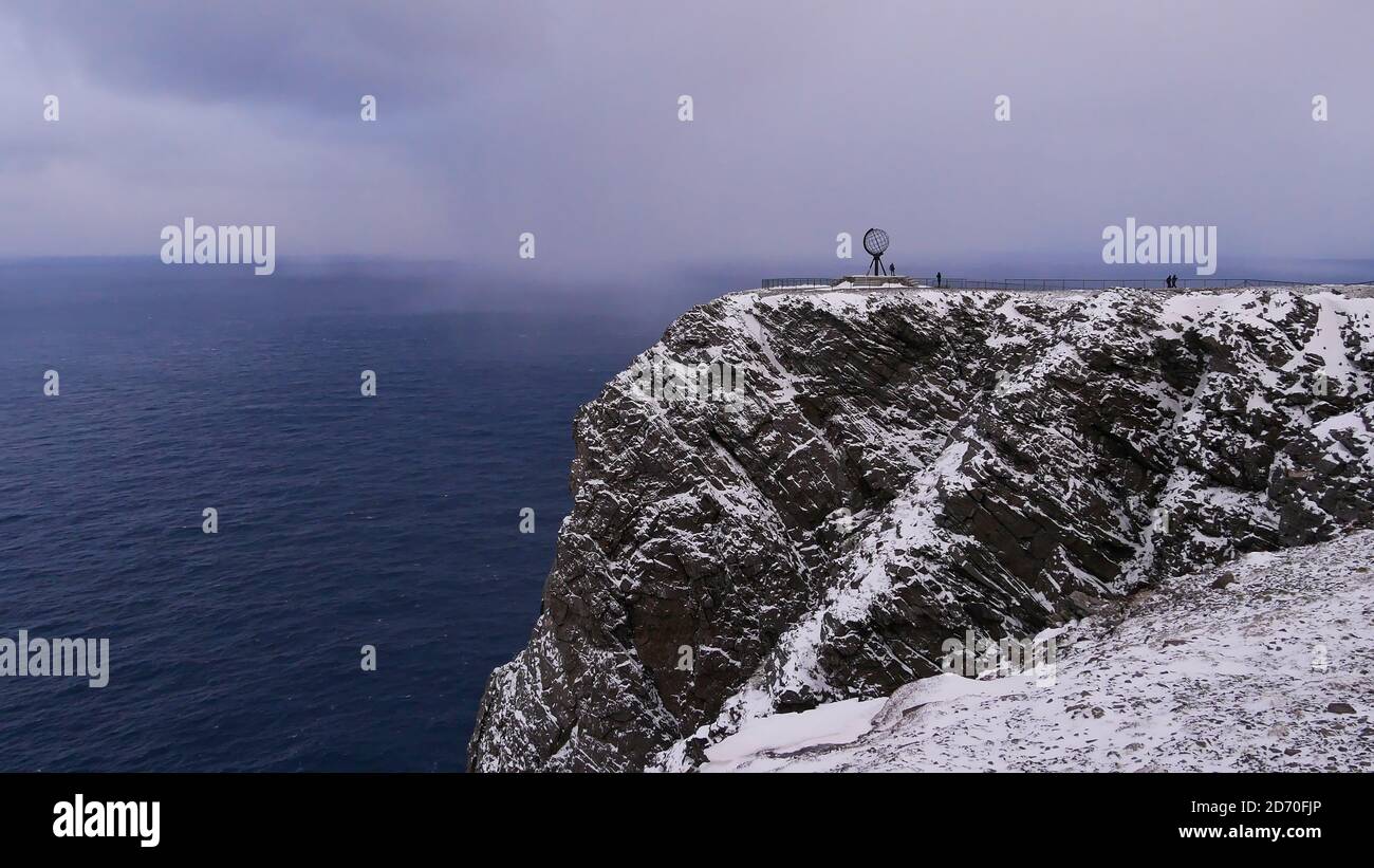 Sculpture populaire de globe sur une falaise rocheuse enneigée du Cap Nord (Nordkapp), en Norvège, en Scandinavie avec peu de touristes et une mer arctique rugueuse en hiver. Banque D'Images