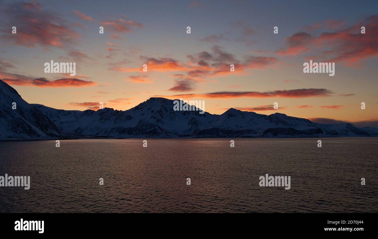 Vue panoramique stupéfiante après le coucher du soleil sur les montagnes enneigées de la côte norvégienne, la Scandinavie avec des nuages orange reflétés dans l'eau. Banque D'Images
