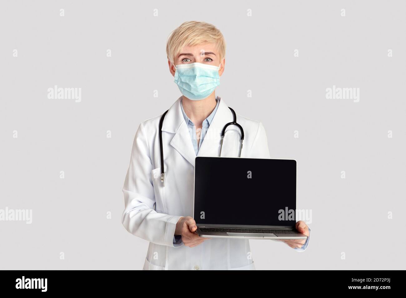 Femme adulte sérieuse en manteau blanc avec stéthoscope et protecteur le masque affiche l'ordinateur portable avec un écran vide Banque D'Images