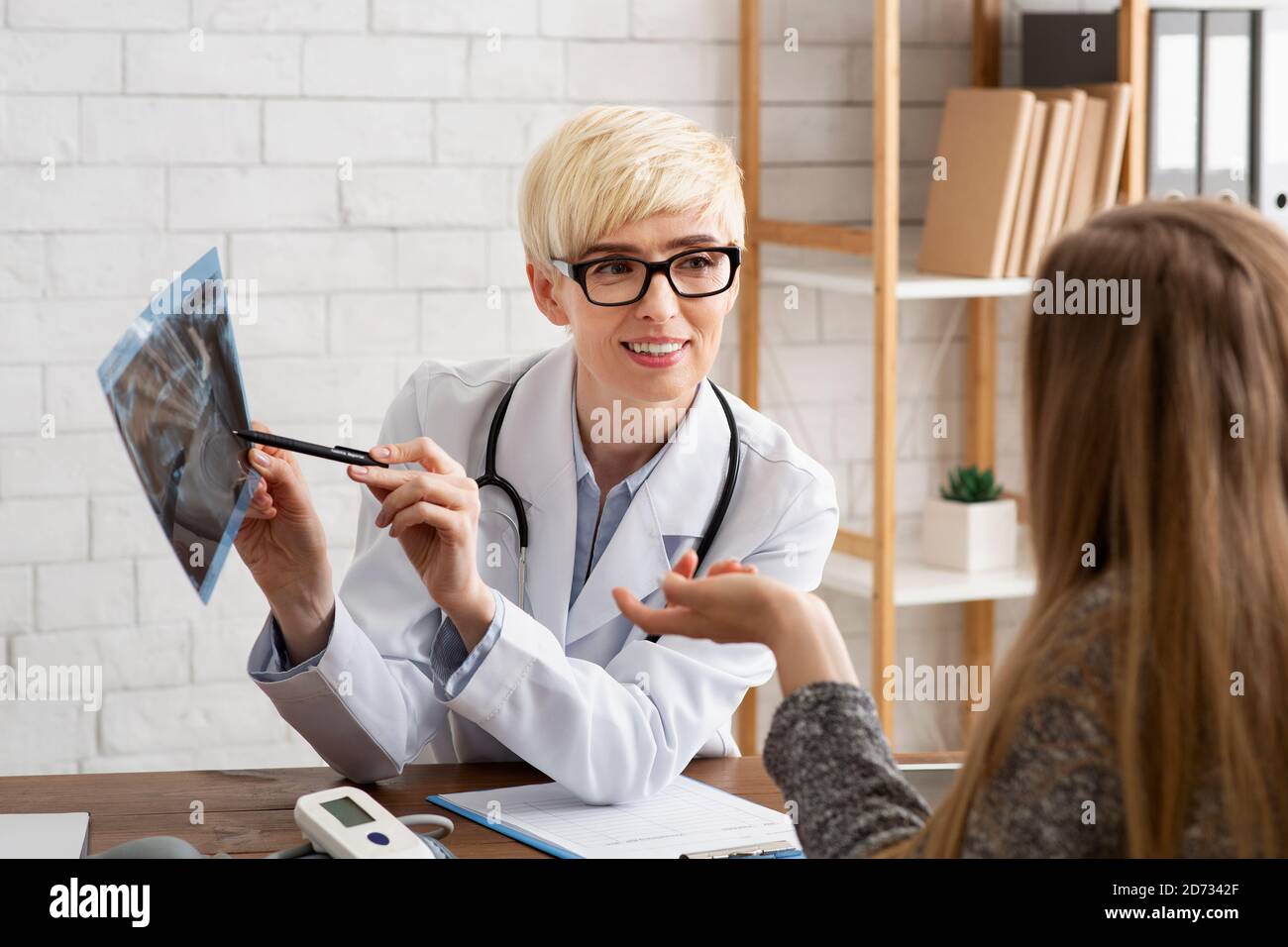 Femme médicale d'âge moyen souriante en lunettes et manteau blanc pointe vers les rayons x et communique avec le patient Banque D'Images
