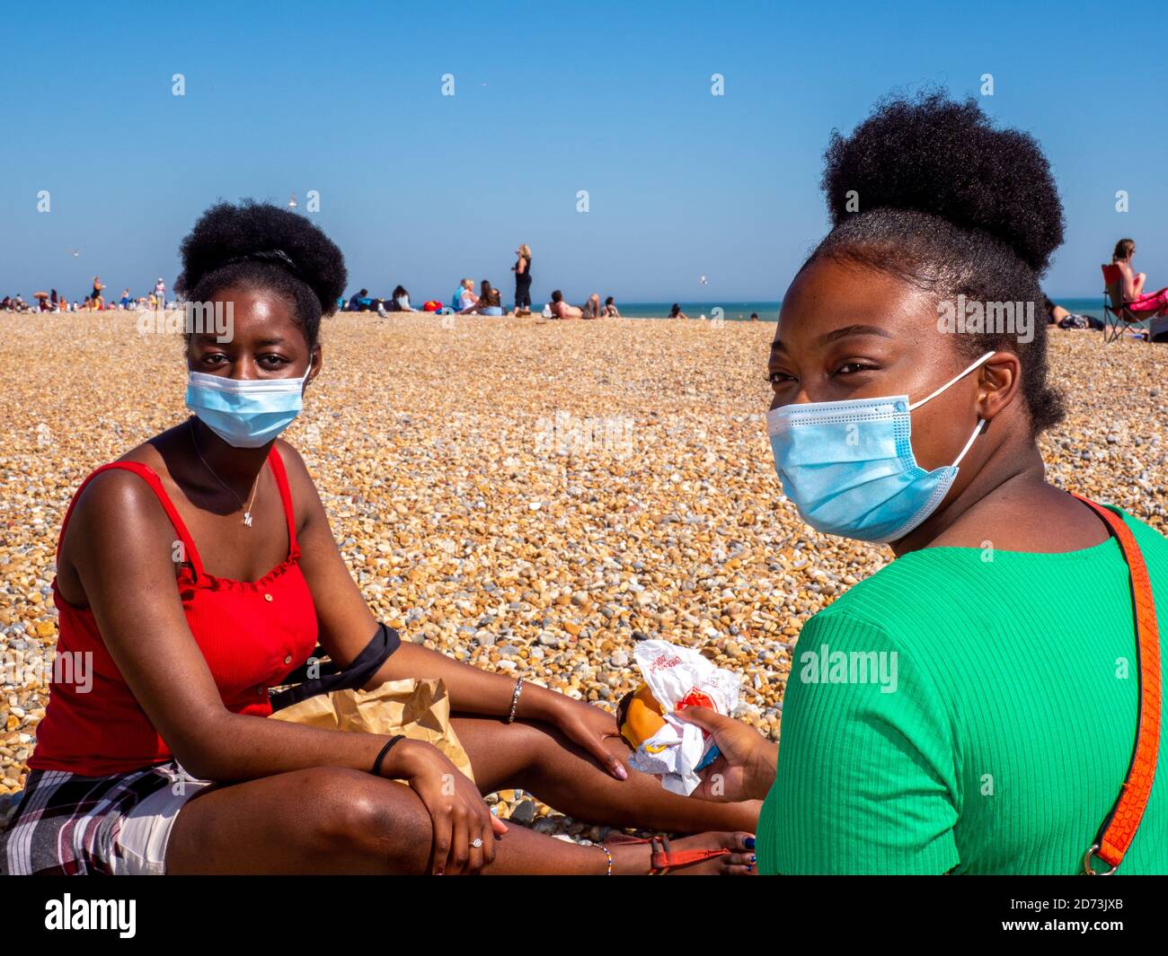 Des amis portant un visage couvrant s'assoient sur la plage de Hastings pour manger un McDonalds. Banque D'Images