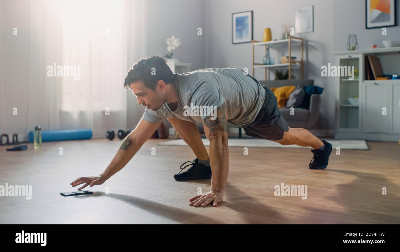 L'homme en T-shirt et short d'athlétisme musculaire fait des exercices de Climber tout en utilisant un chronomètre sur son téléphone. Il est formation à la maison dans Banque D'Images