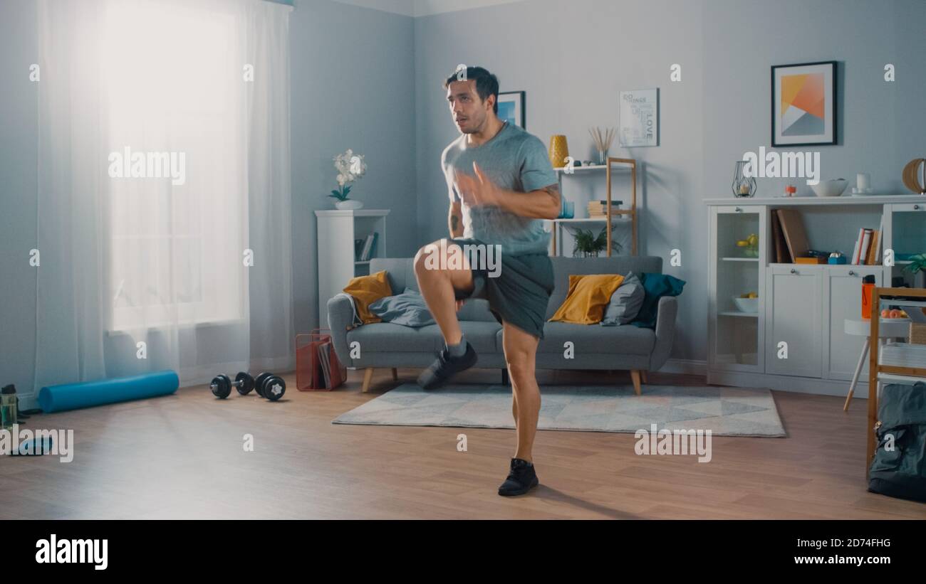 Le modèle Strong Athletic Fit Man en T-shirt et short est énergique Jogging en place à la maison dans son spacieux et lumineux Séjour avec minimalistes Banque D'Images
