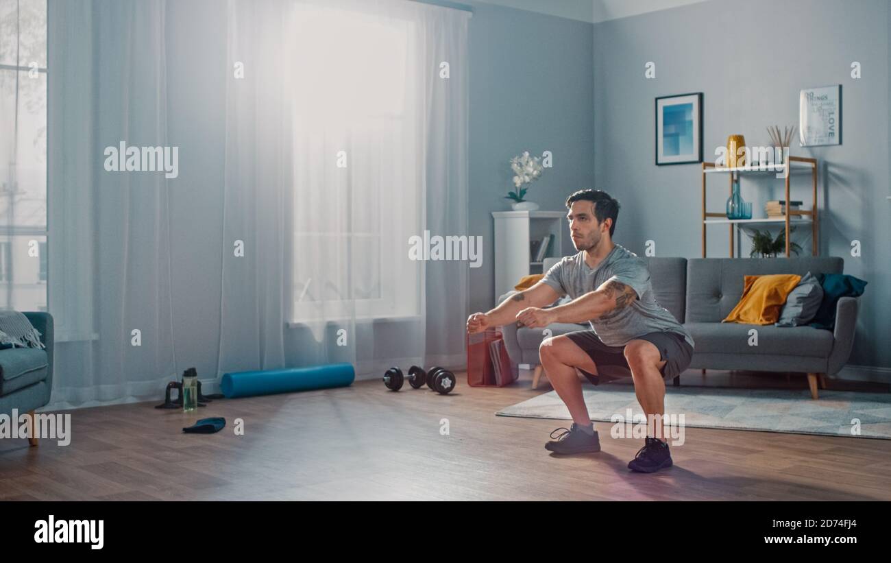 Muscle Athletic Fit Man en T-shirt et short fait des exercices de squat à la maison dans son salon spacieux et lumineux avec intérieur minimaliste. Banque D'Images