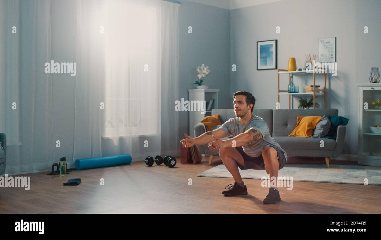 Muscle Athletic Fit Man en T-shirt et short fait des exercices de squat à la maison dans son appartement spacieux et lumineux avec intérieur minimaliste. Banque D'Images