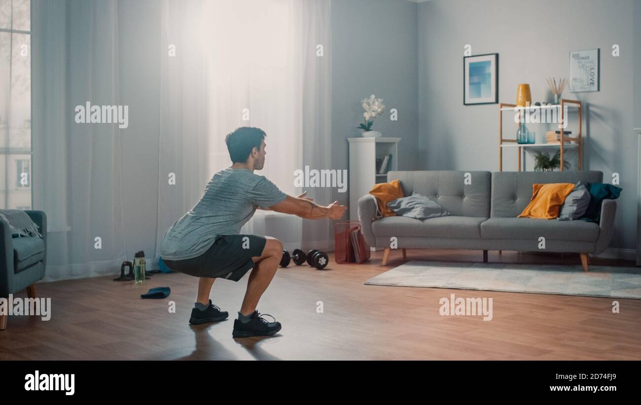 L'homme puissant Athletic Fit en T-shirt et short fait des exercices de squat à la maison dans son appartement spacieux et lumineux avec intérieur minimaliste. Banque D'Images