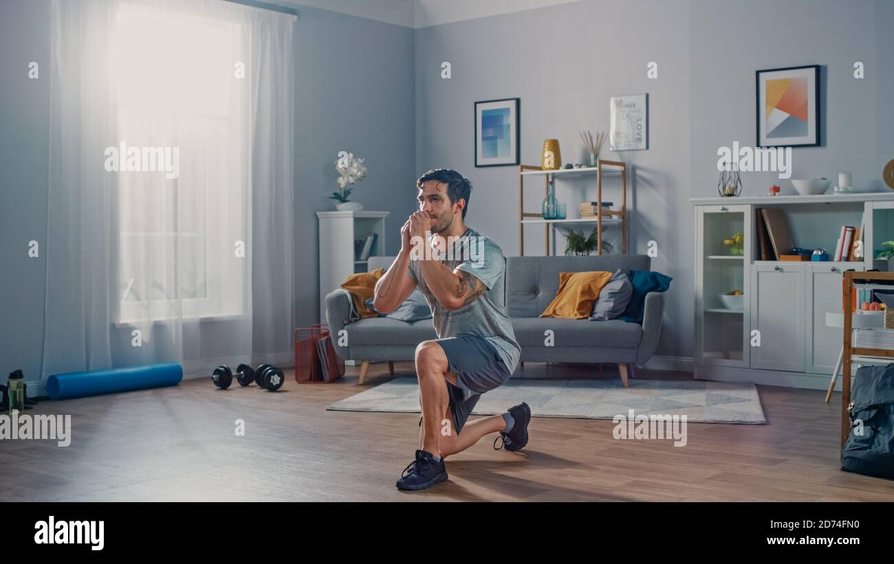 Un homme puissant et athlétique en T-shirt et en short est en train de faire Exercices de fente avant à la maison dans son spacieux et lumineux Appartement avec intérieur minimaliste Banque D'Images
