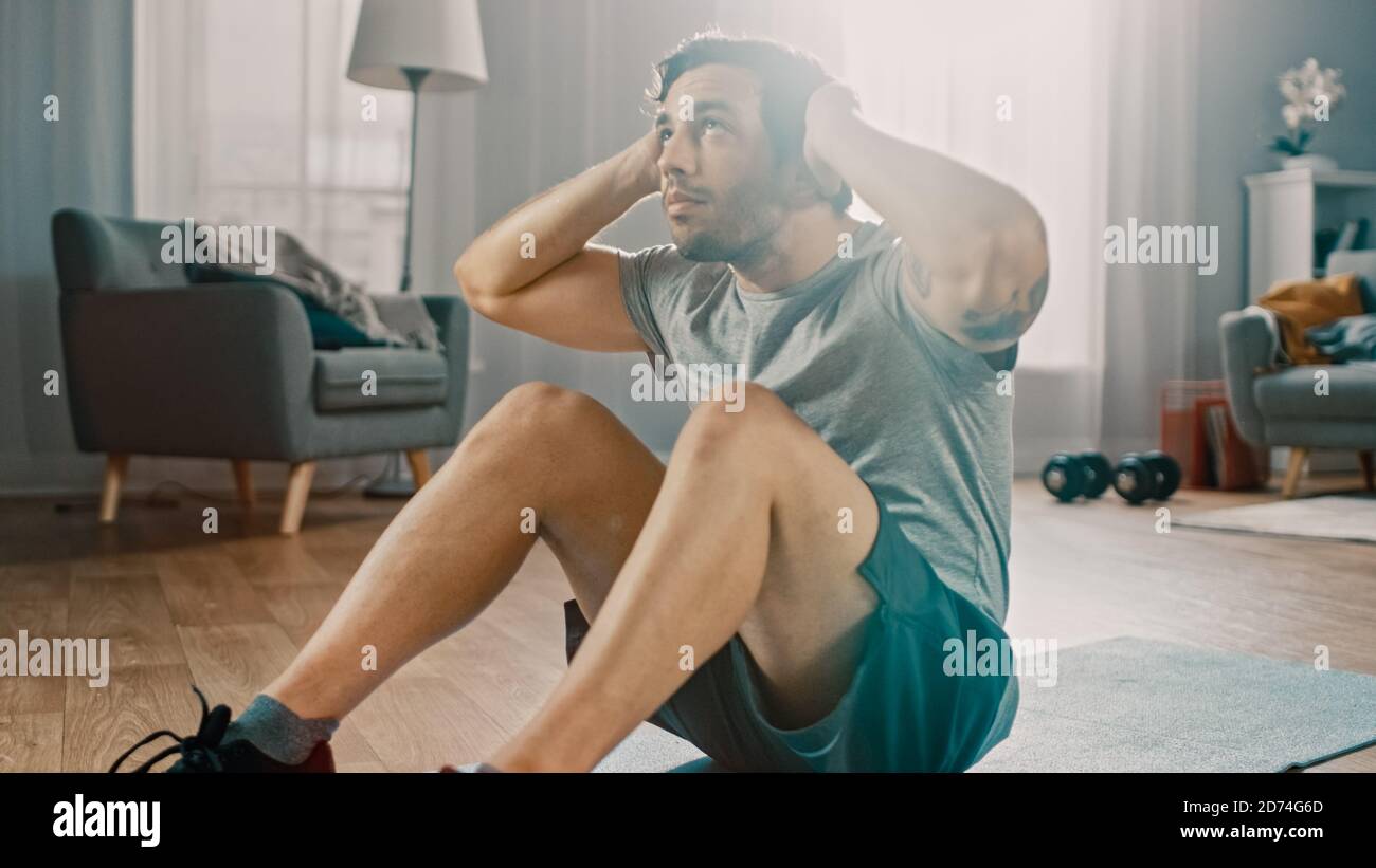 Un homme puissant et athlétique en T-shirt et en short est en train de faire Abdominal Crunch s'entraînement à la maison dans son spacieux et lumineux Séjour avec minimalistes Banque D'Images