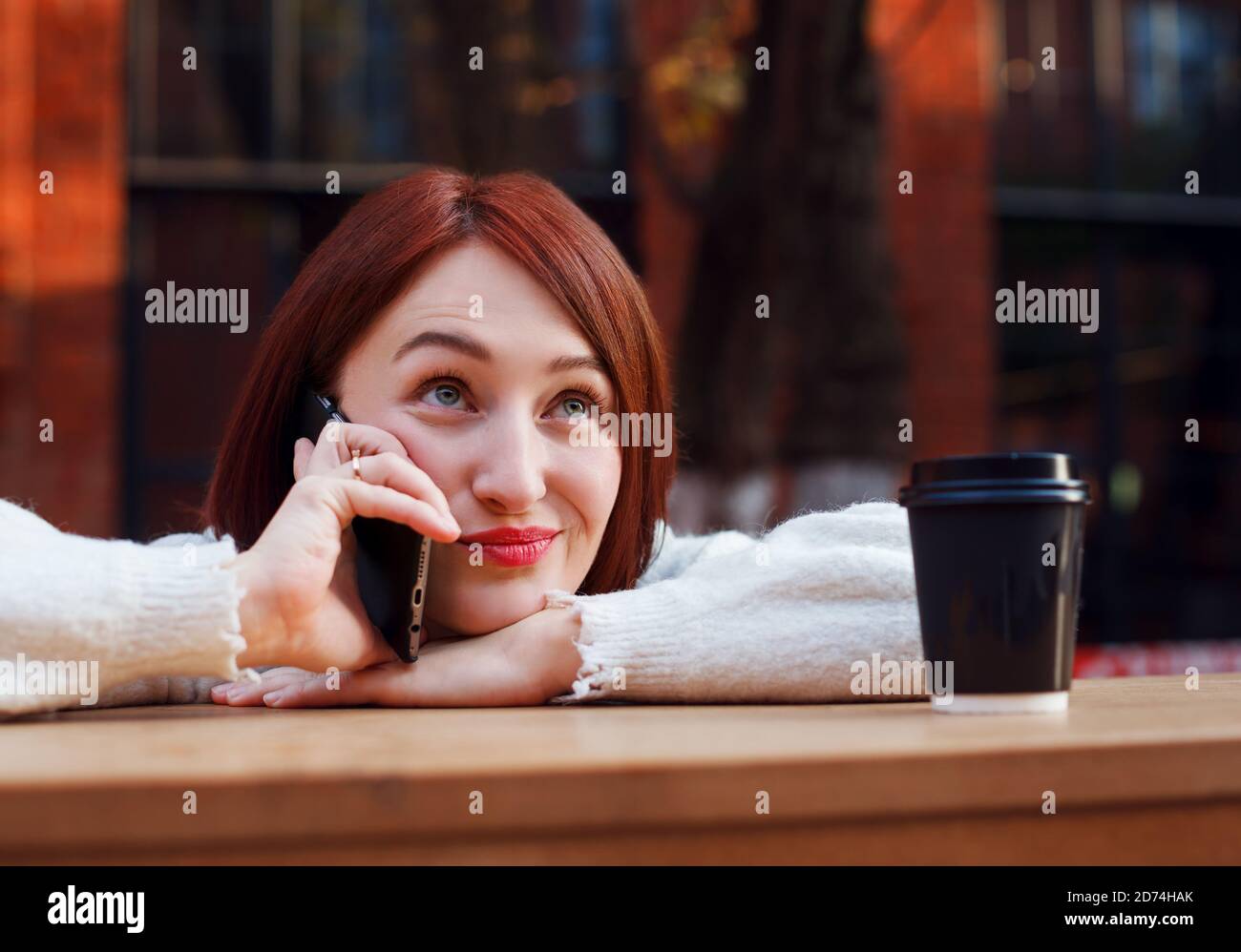Elle s'appuie sur une table de bar sur la terrasse d'un café en plein air et parle au téléphone avec une drôle d'expression émotionnelle sur son visage. Banque D'Images