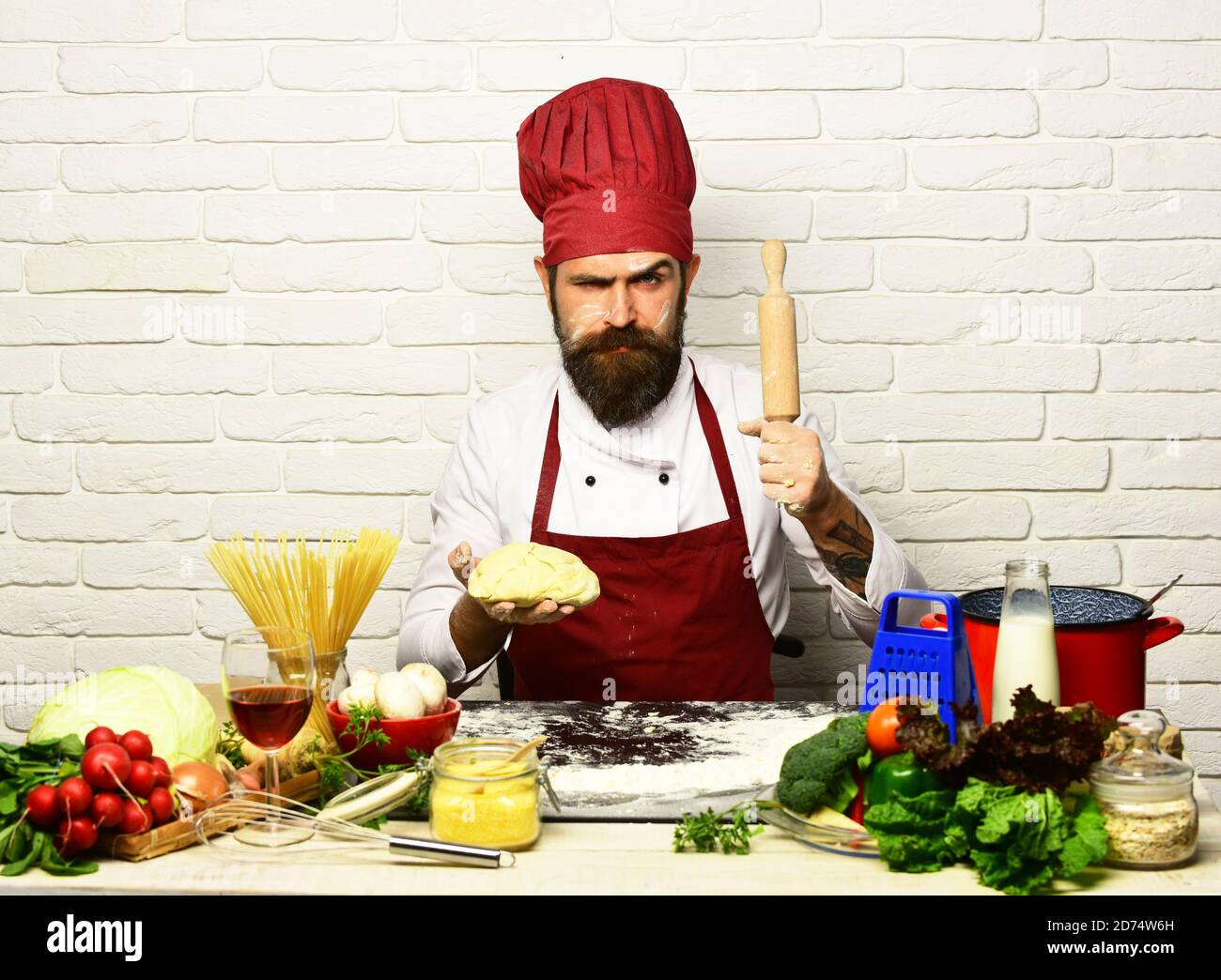 Le chef prépare de la pâte. Concept de cuisine italienne. Homme avec barbe  tient la pâte et la broche sur fond de brique blanche. Cuire avec le visage  strict en uniforme bordeaux