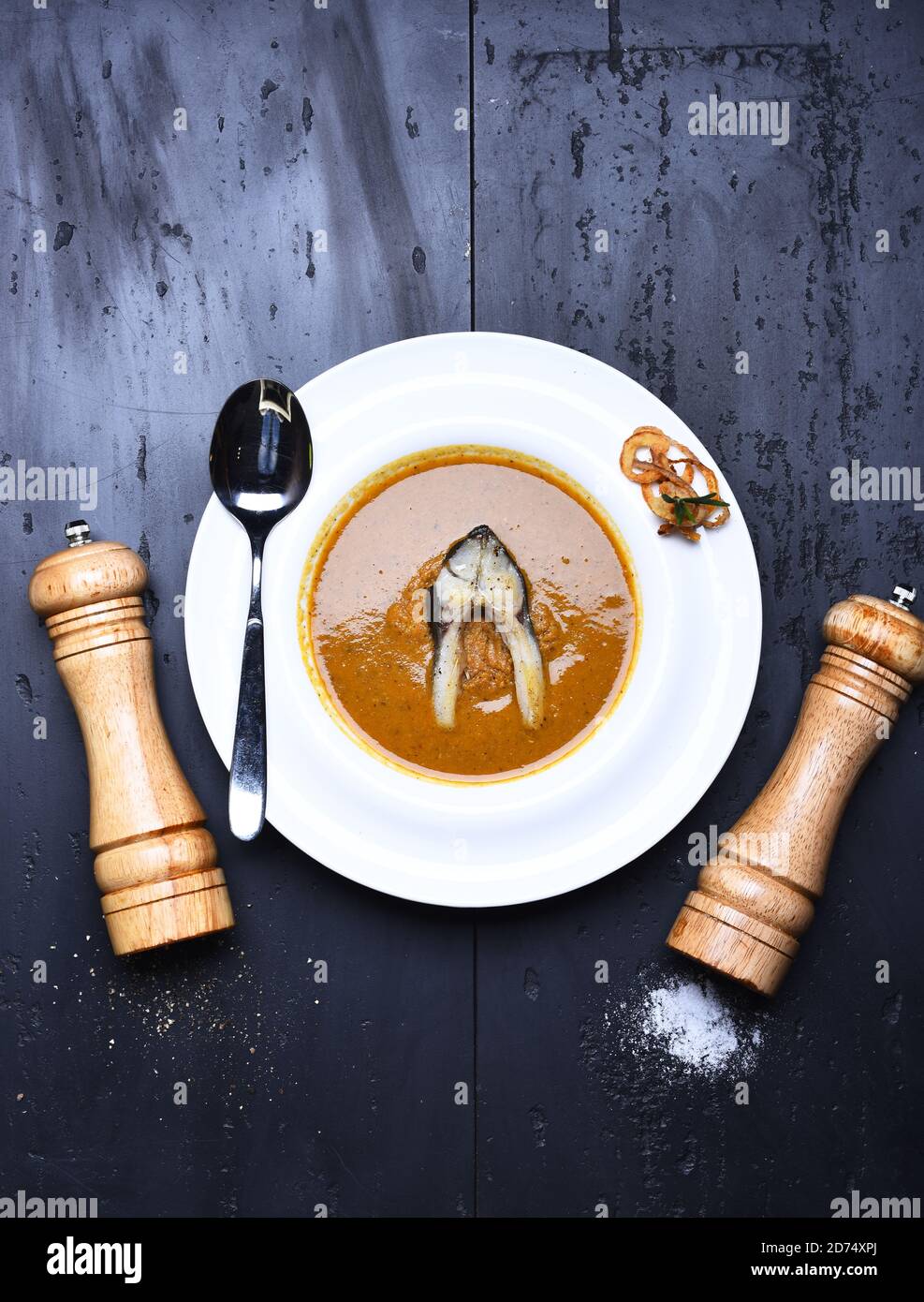 Poivrière en bois, saltcave et cuillère près du plat. Concept de cuisine française. Soupe à la crème ou purée brune sur fond de texture grise. Soupe de poisson avec un morceau de poisson et des rondelles d'oignon frits sur une assiette blanche Banque D'Images