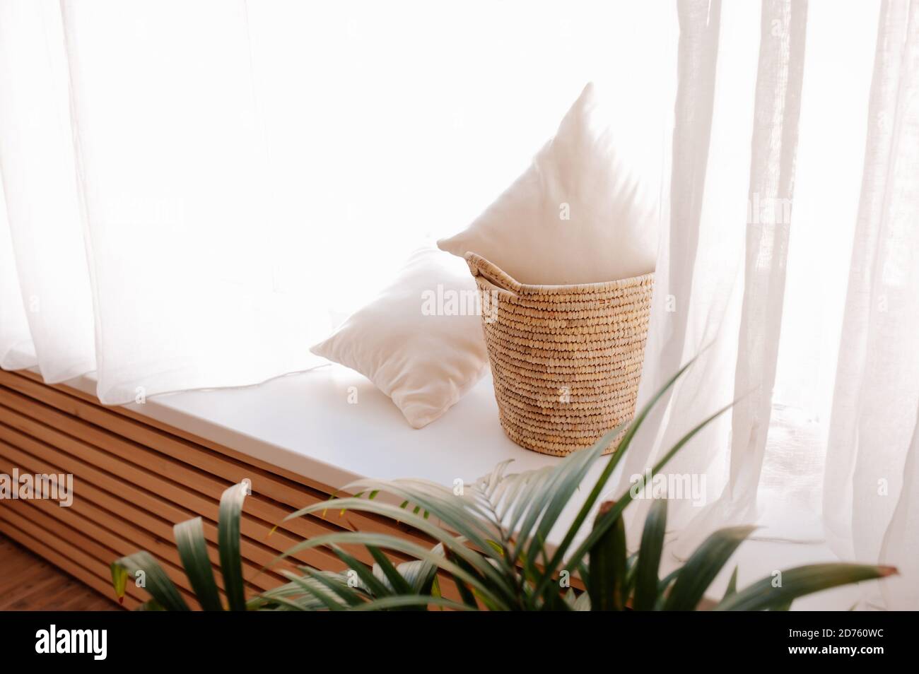 Oreillers doux et légers dans un grand panier en osier sur la fenêtre avec rideaux blancs et feuilles de palmier. Style scandinave minimaliste. Rangement confortable et pratique Banque D'Images