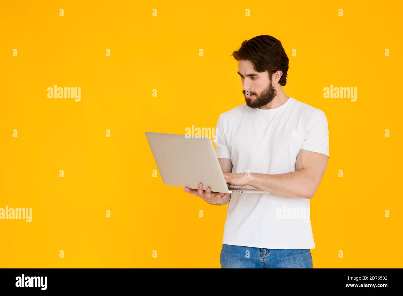 Un jeune homme barbu sur fond jaune avec un l'ordinateur portable entre ses mains regarde l'écran et sourit Banque D'Images