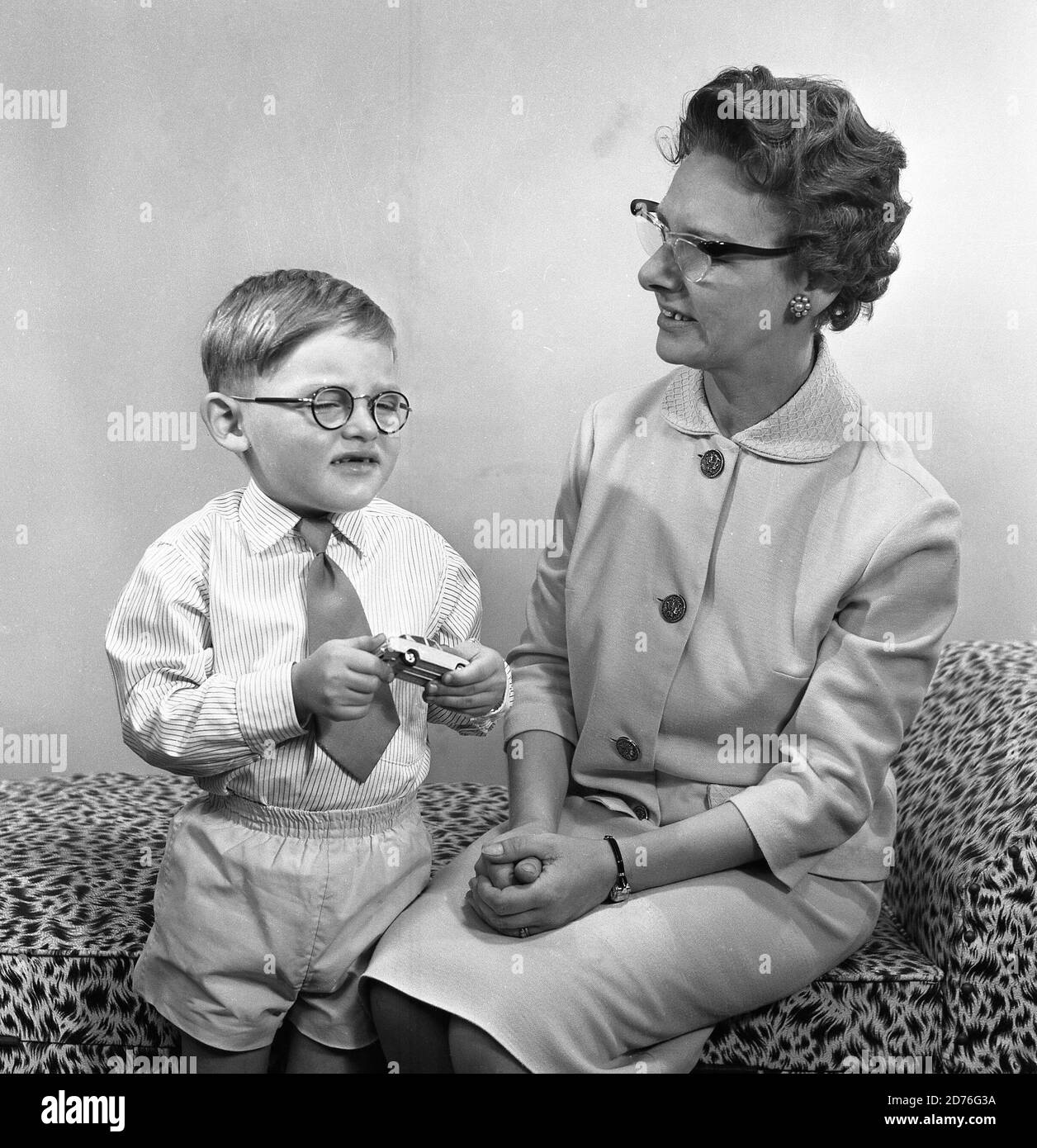 années 1960, historique, comme la mère, comme le fils! Petit garçon avec de petites lunettes rondes et tenant sa voiture en métal jouet dans les mains debout à côté de sa mère, qui porte également une paire de lunettes, qui ont été l'un des styles féminins populaires cette époque, Angleterre, Royaume-Uni. Banque D'Images