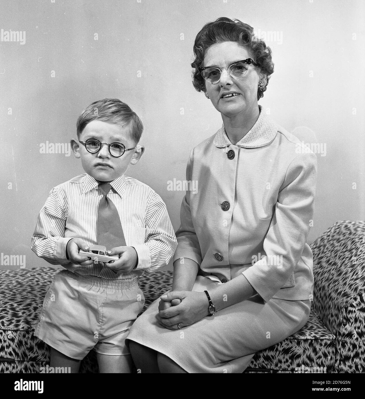 années 1960, historique, comme la mère, comme le fils! Petit garçon avec de petites  lunettes rondes et tenant sa voiture en métal jouet dans les mains debout à  côté de sa mère,