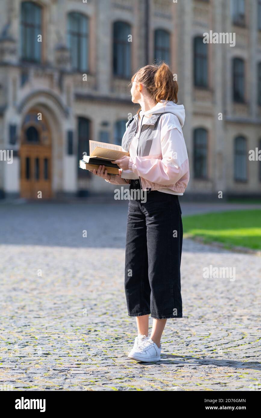 Jeune femme tendance portant des livres dans un quadrilatère ou urbain parc debout avec vue sur une épaule à un historique bâtiment Banque D'Images