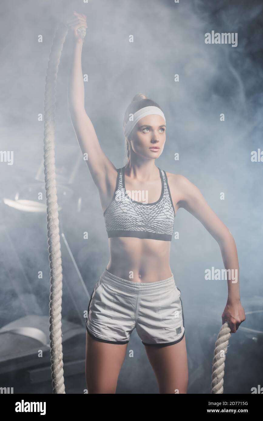 Objectif sélectif de la jeune sportswoman tenant la corde de bataille dans la salle de gym avec fumée Banque D'Images