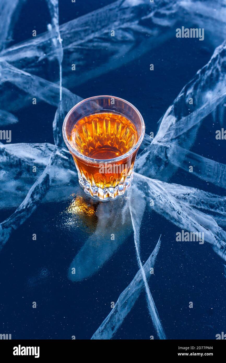 Un verre de whisky se trouve sur une glace aux fissures profondes. Liquide orange sur fond bleu. Vue de dessus de côté. Banque D'Images