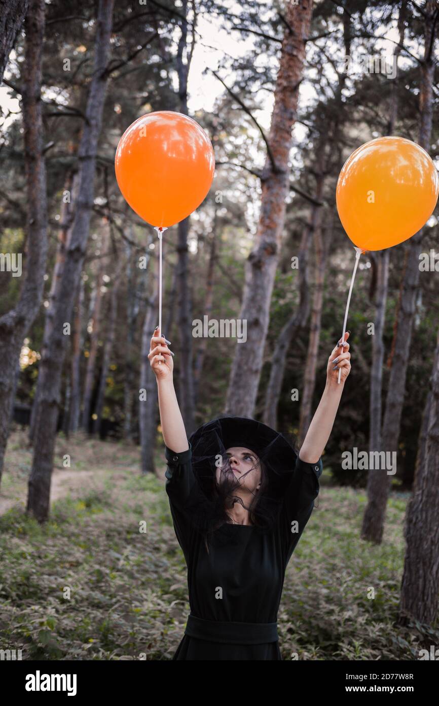 Jeune mignonne belle femme en robe sombre et chapeau de sorcière tient des ballons d'orange dans ses mains. Costume de fête d'Halloween. Forêt, parc avec arbres d'automne Banque D'Images