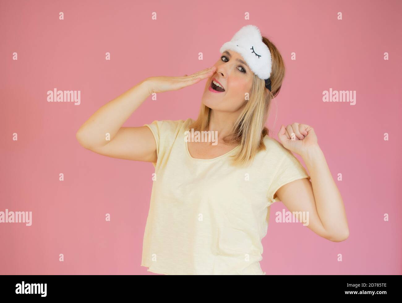 Jeune femme fatiguée en pyjama jaune maison porter un masque de sommeil bâilling couvrant la bouche avec la main reposant à la maison isolée sur fond rose pastel studio po Banque D'Images