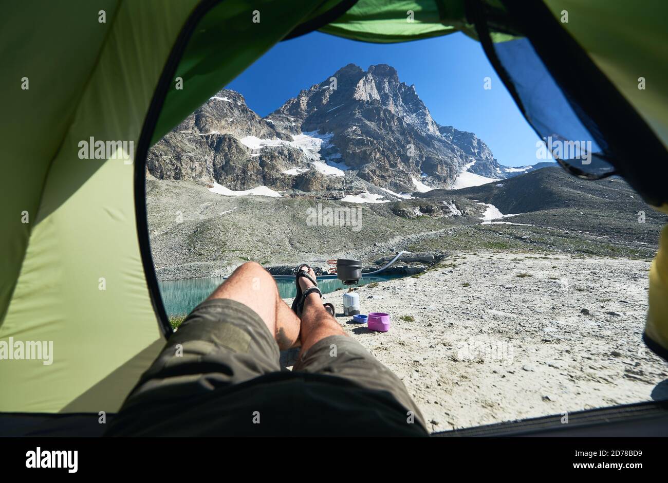 Vue depuis la tente sur le lac de montagne avec eau claire et fraîche, jambes de mâles de touristes incognito et de l'équipement dans la journée ensoleillée d'été. Attraction populaire dans les Alpes rocheuses. Concept de tourisme, camping, beauté de la nature. Banque D'Images