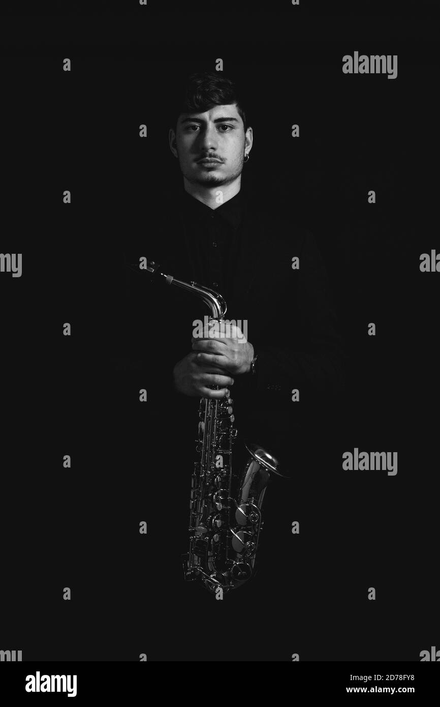 Prise de vue en niveaux de gris d'un gars sympa et charmant tenant son saxophone sur fond sombre Banque D'Images
