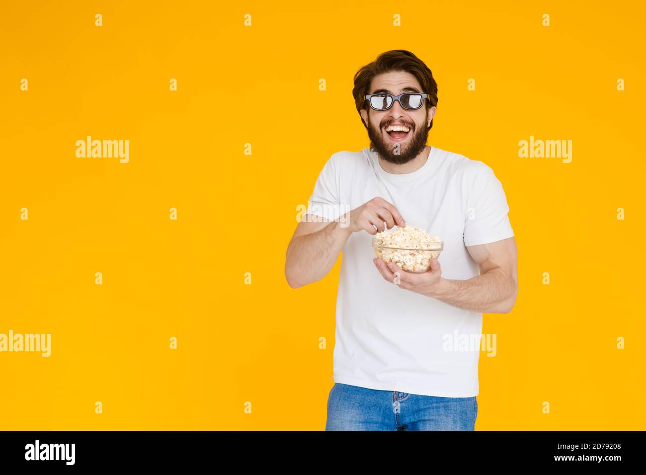 Image d'un rire heureux jeune homme en t-shirt blanc manger du popcorn portant des lunettes 3d voir les films plus isolé sur fond jaune. Banque D'Images