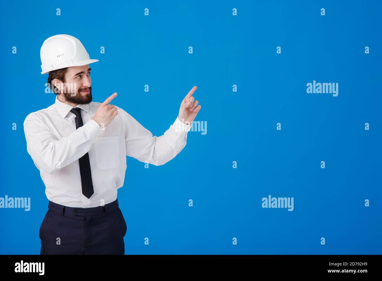 Beau homme ingénieur industriel portant un casque blanc et pointant le doigt vers un espace de copie vierge pour le texte isolé sur bleu Banque D'Images