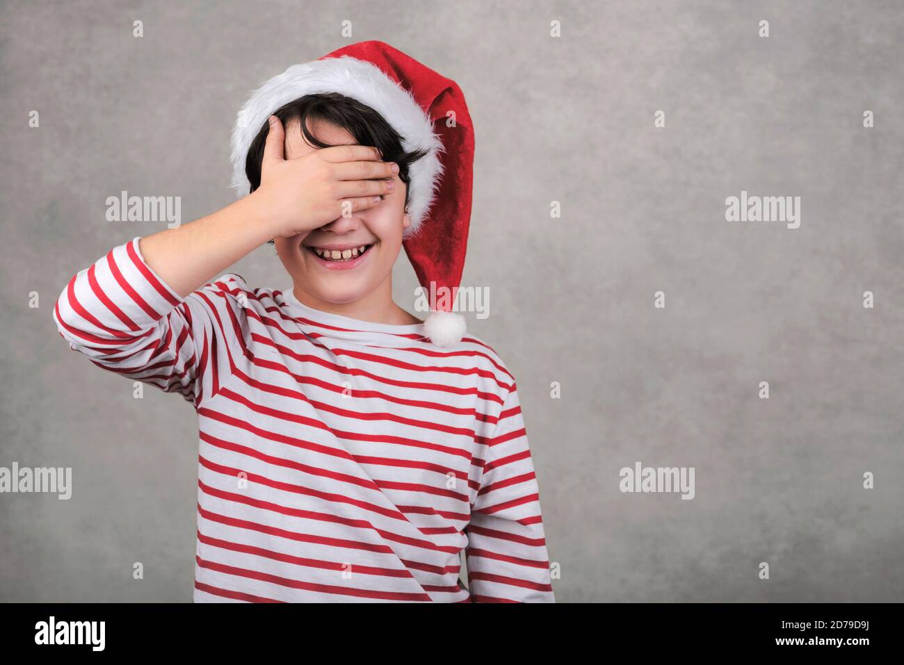 Joyeux Noël, jeune enfant souriant portant un chapeau de Père Noël couvrant ses yeux avec sa main sur fond gris Banque D'Images