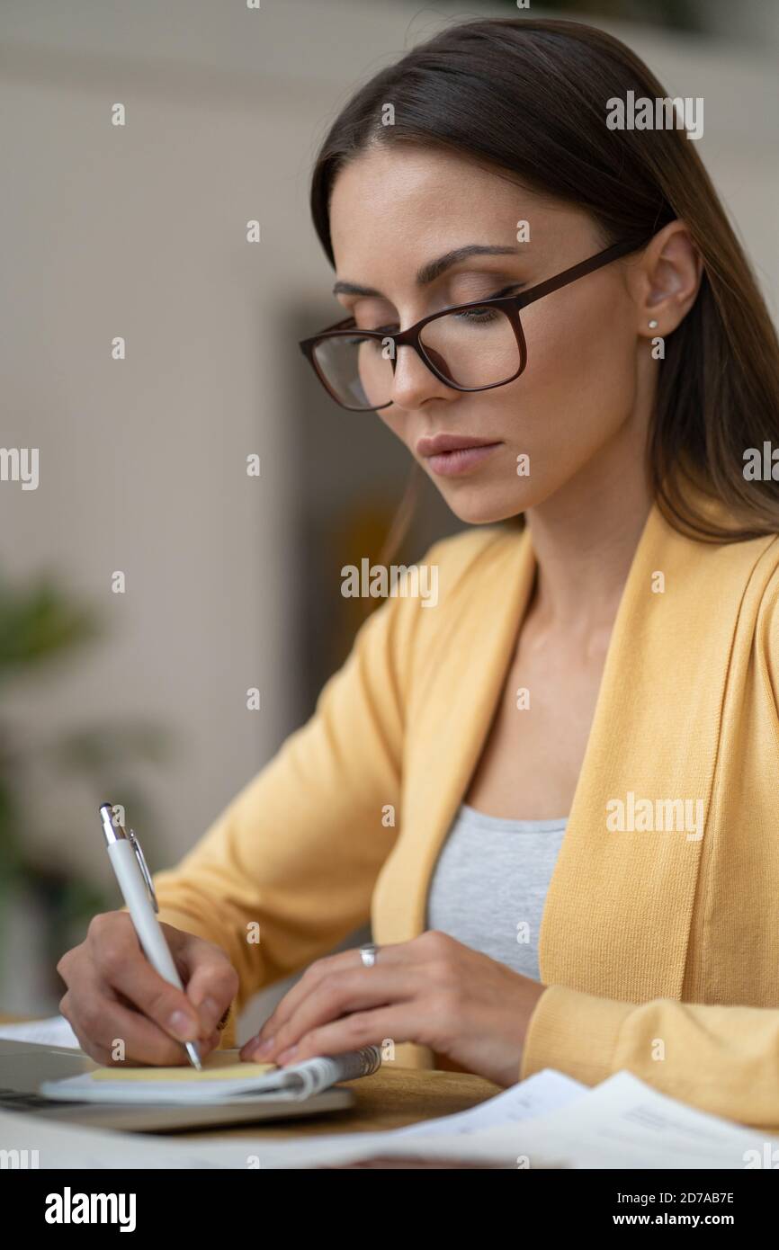 Femme d'affaires ciblée en lunettes porter un gilet jaune pour faire une note dans un carnet. Une femme de bureau travaille à distance depuis la maison pendant la seconde Banque D'Images