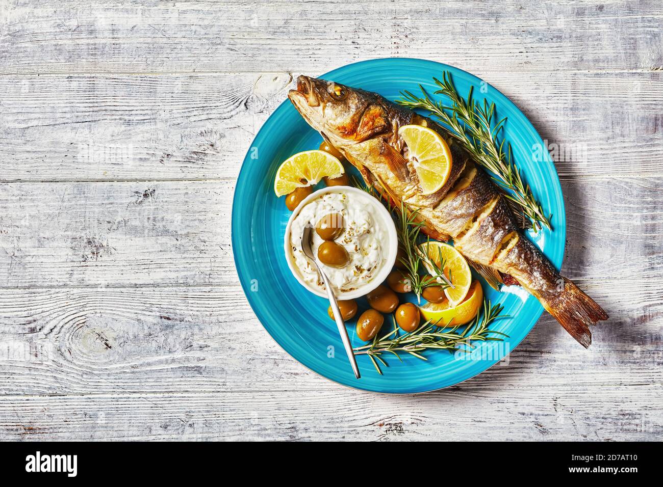 Poisson de mer rôti entier avec des quartiers de citron, du romarin, du persil, servi sur une assiette bleue sur un fond de bois avec des olives vertes coutellerie et du tartre sa Banque D'Images