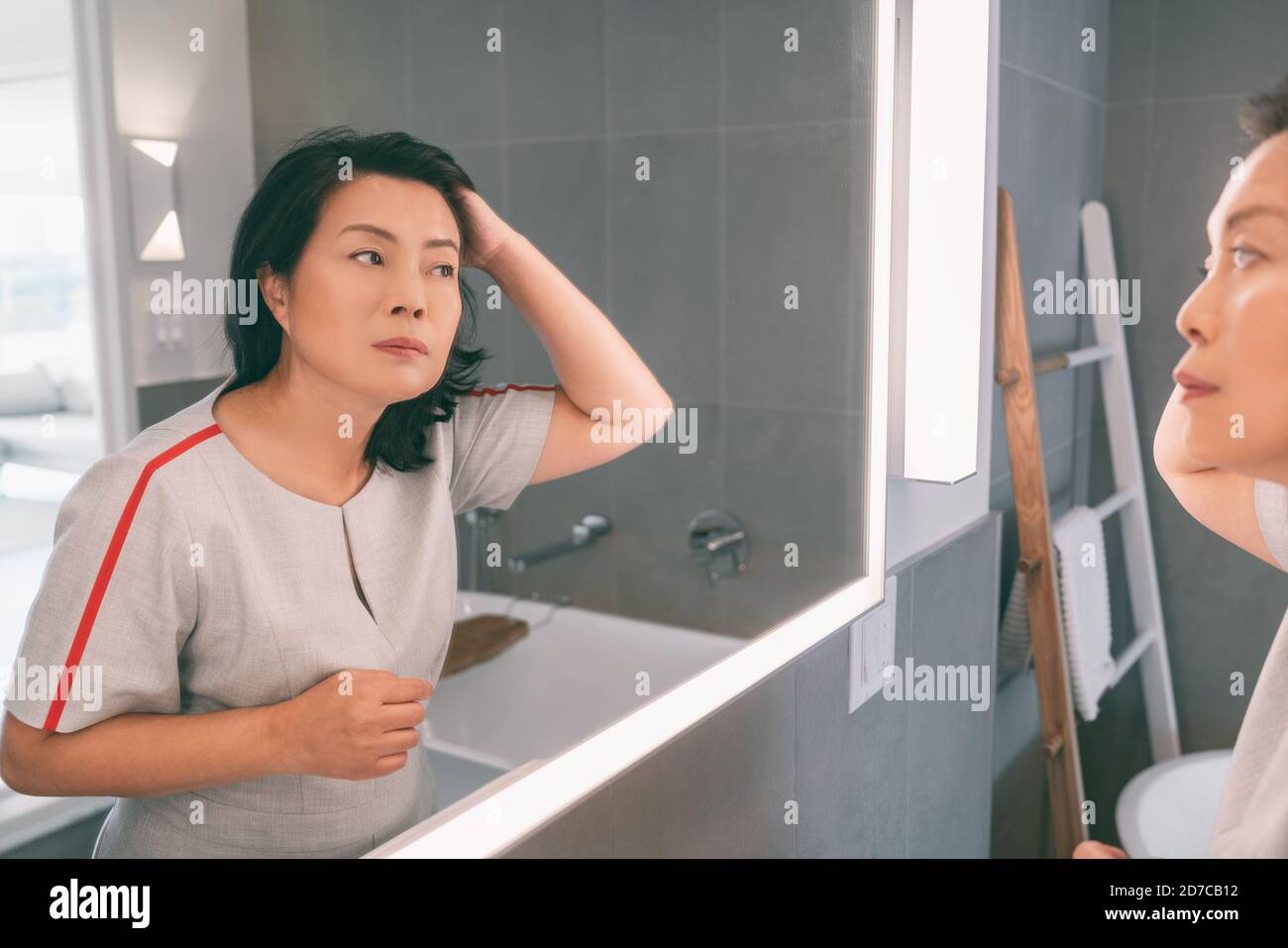 Perte de cheveux mature femme asiatique touchant ses cheveux coiffant ou colorant les cheveux gris regardant elle-même dans le miroir de la maison. Belle dame chinoise en elle Banque D'Images