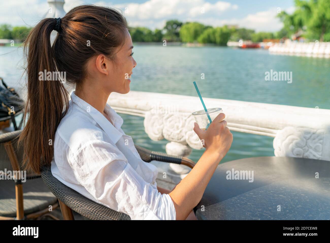 Bonne jeune femme asiatique se détendant au café du port en bord de mer pour boire boisson fraîche au café en profitant de la journée d'été dans la ville par le lac Banque D'Images
