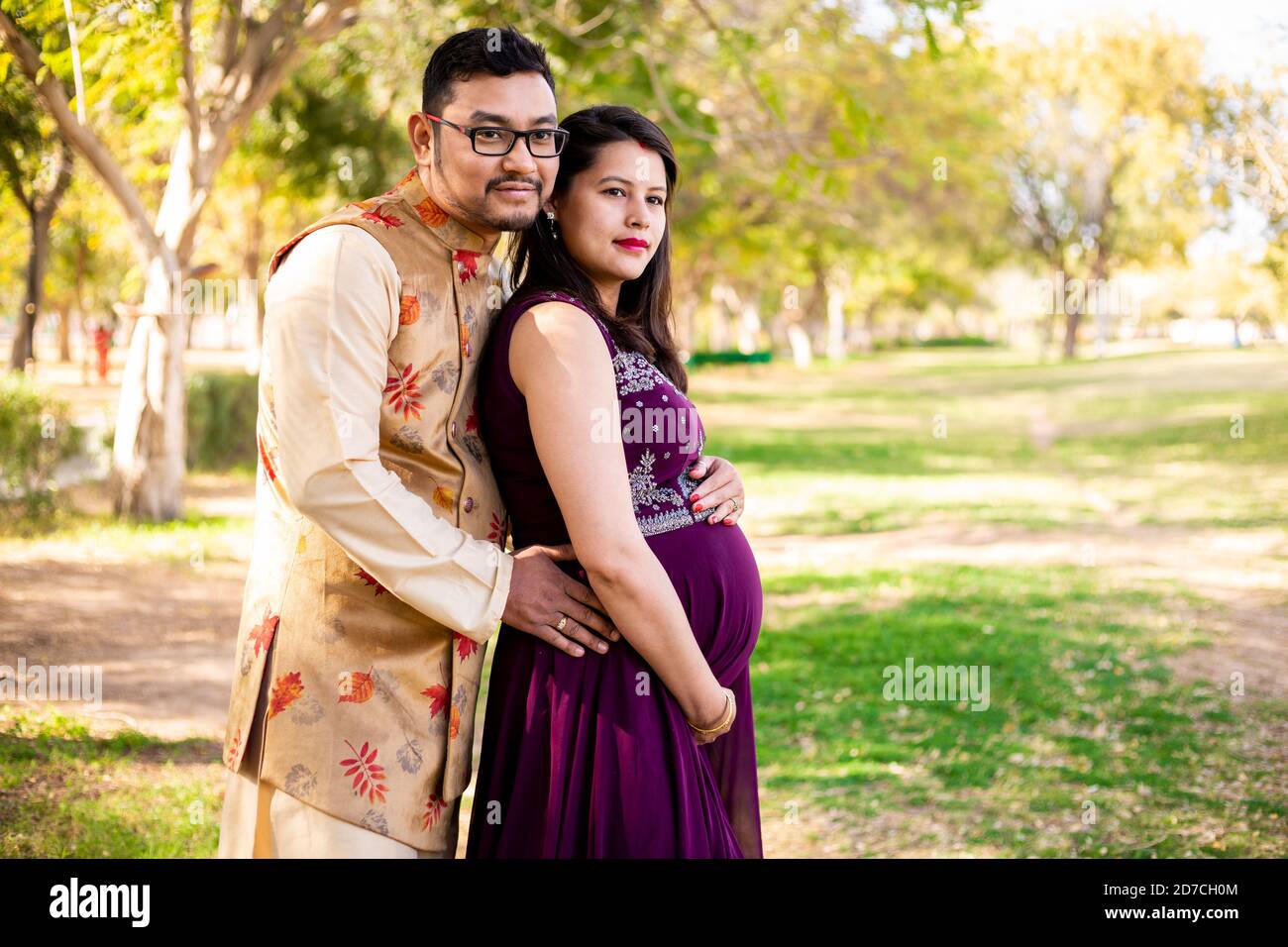Jeune femme enceinte asiatique indienne avec son mari portant une tenue traditionnelle debout dans un parc ou un jardin pour écrire du texte. Banque D'Images
