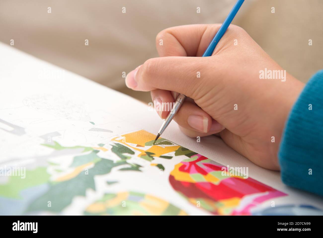 8 - gros plan détaillé d'un pinceau fin utilisé par une jeune femme pour peindre par des nombres. Plusieurs couleurs et des bleus éclatants contrastent la peau chaude. Banque D'Images