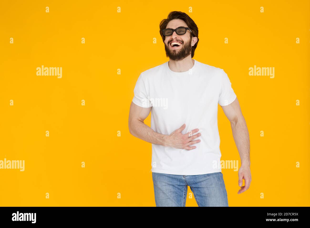 Rire jeune barbu homme gars en t-shirt blanc 3d lunettes isolées sur fond jaune. Concept de mode de vie des personnes. Regarder un film , mettez la main sur Banque D'Images