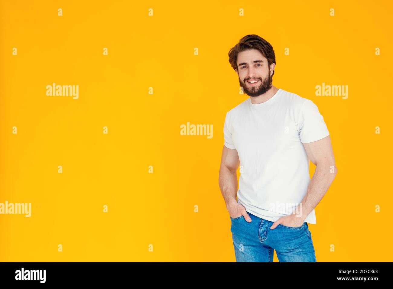un jeune homme barbu des années 30 sur fond jaune. un homme joyeux et joyeux dans un t-shirt blanc et un pantalon en denim sourit à l'appareil photo debout Banque D'Images