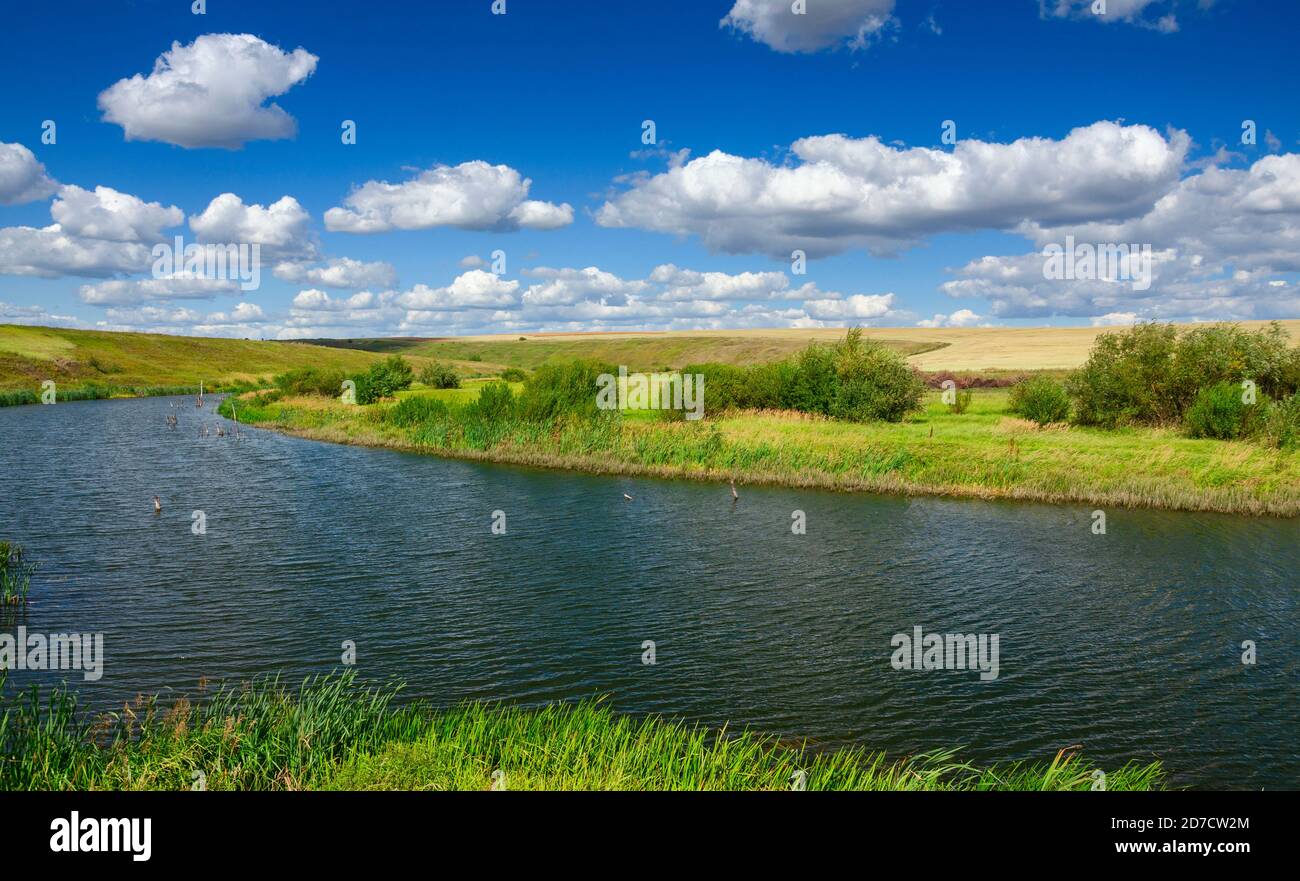 Paysage ensoleillé d'été avec rivière, champs, collines vertes et de beaux nuages dans le ciel bleu. Banque D'Images