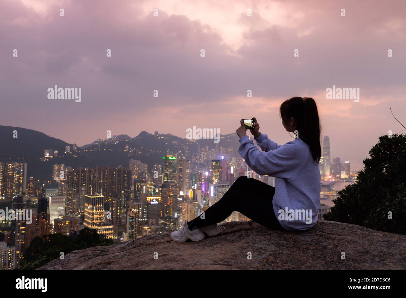 Silhouette de fille qui s'assoit au rocher et prend des photos de la vue de nuit de la ville Skyline de l'île de Hong Kong (au sud du port de Victoria) de la colline de Braemar, Banque D'Images