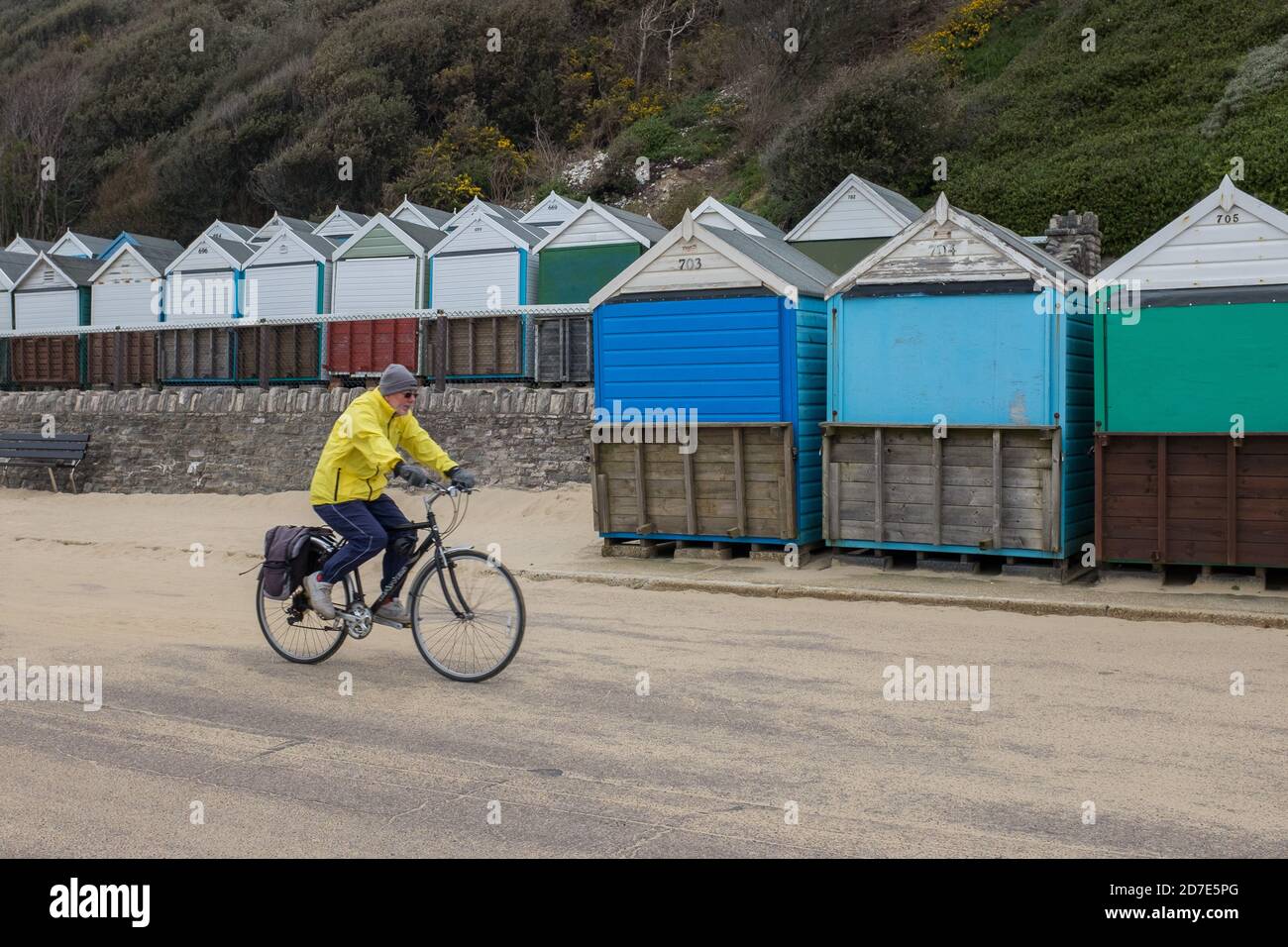 Un cycliste de couleur jaune vif, sous un manteau coupe-vent, passe devant des rangées de cabanes de plage le long de la promenade couverte de sable soufflé sur la plage de Boscombe à Bournemouth le jour de février. 10 février 2015. Photo: Neil Turner Banque D'Images