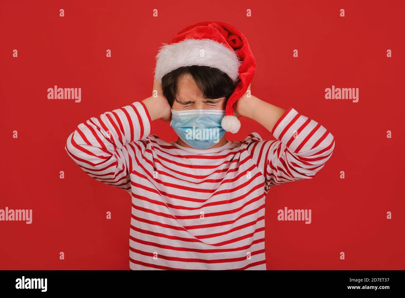 Enfant en colère avec un masque médical portant un chapeau de Père Noël avec ses mains sur la tête sur fond rouge Banque D'Images