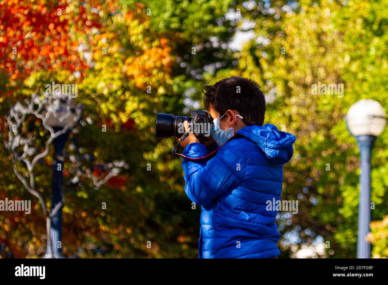 Un garçon caucasien portant un masque facial en raison de la pandémie de COVID-19 prend une photo du paysage d'automne à l'aide d'un appareil photo reflex numérique professionnel. Arbres colorés Banque D'Images