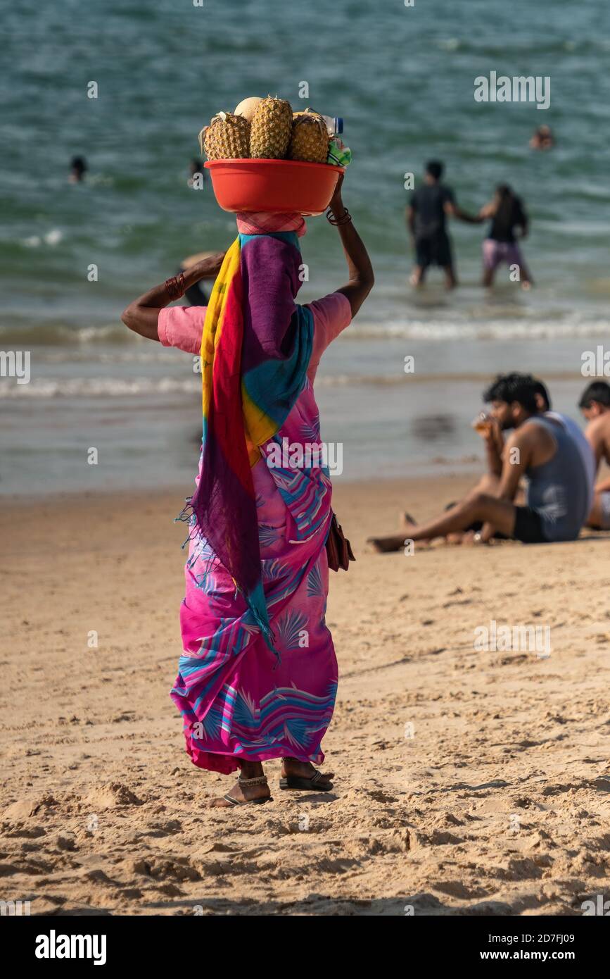 Femme indienne non identifiée en rose coloré bleu sari robe portant des fruits frais dans le bassin rouge à sa tête pour vendre à la rive aux touristes. Ensoleillé Banque D'Images