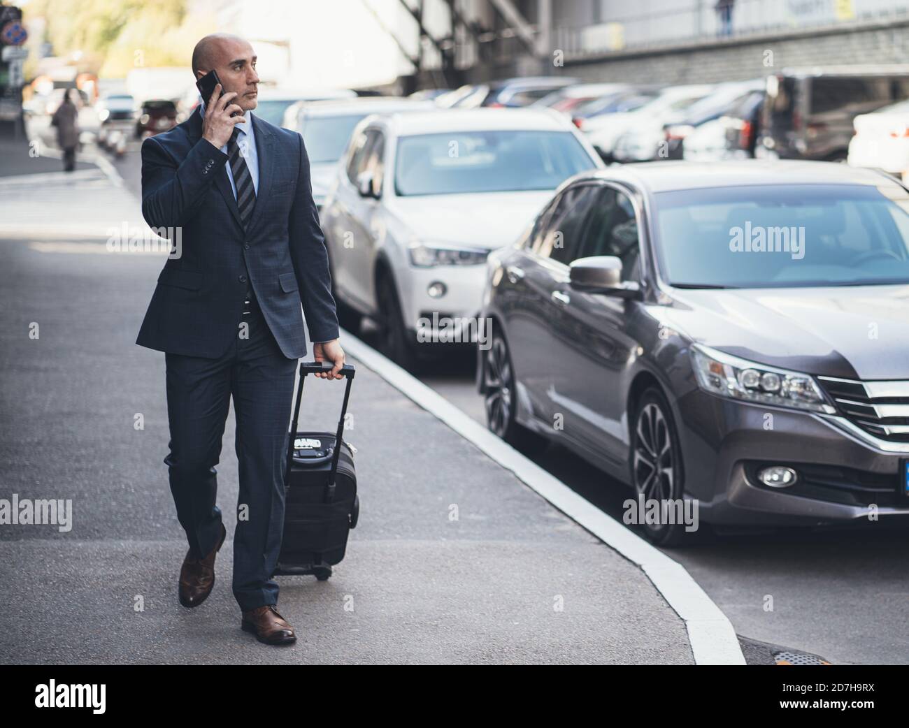 Homme d'affaires parle sur téléphone portable tenant une valise à la main. Homme caucasien d'âge moyen marchant le long du trottoir devant les voitures garées. Concept de voyage d'affaires Banque D'Images